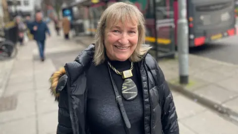 Carol Murphy is smiling. She in a city centre. She's wearing a necklace large with a large pendant and a black coat. A bus is behind her and people are in the background walking. 