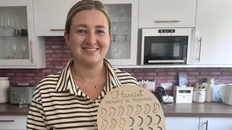 A woman with light brown hair, tied back, stands in a kitchen while holding a wooden midwife-in-training feet counter, with features foot-shaped spaces for miniature wooden feet to be inserted, along with the words: "Harriet, midwife in training." She is wearing a black and cream polo shirt.