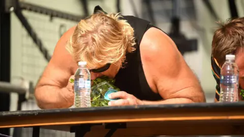 Glenn Walsh in a black vest and sunglasses leaning over a table and pushing watercress into his mouth