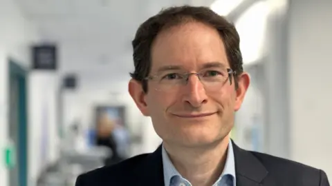 A man with glasses and dark hair is looking straight at the camera in this close-up image. He is wearing a dark jacket and light blue shirt and the background, what could be a corridor, is blurred behind him.