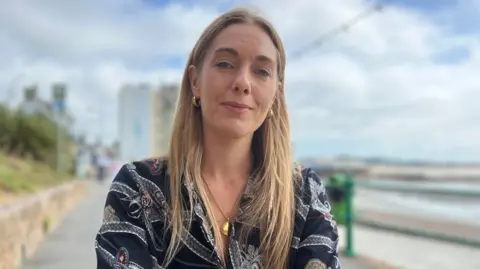 Jessica Pinel in a patterned dark shirt sits on a seafront promenade beside a beach. A stone wall lines the walkway on one side, with railings and the shoreline on the other under a cloudy sky.