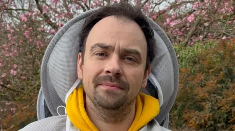 The image shows Dennis Kennedy wearing a protective bee suit with a yellow hoodie sticking out from underneath. Dennis looks expressionlessly at the camera. He has short, dark brown hair and a beard. Bushes and a tree can be seen in the background.