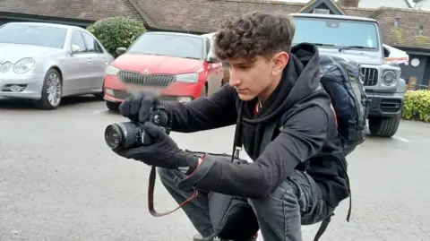 Isaac Potter crouches in a car park as he takes a photo with a camera. He has curly, brown hair and wears grey jeans and a dark hoody.