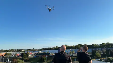 Two men in black shirts standing on the roof of the building and looking up at a small flying drone in a blue sky