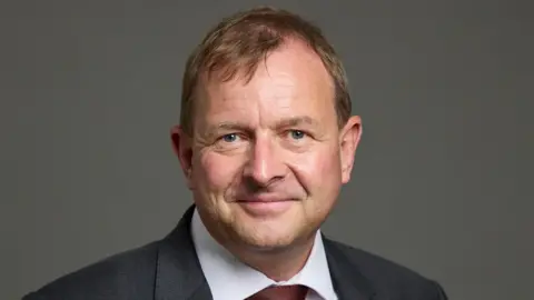 Lord Fuller in a dark jacket, a white shirt and red tie looks straight at the camera 