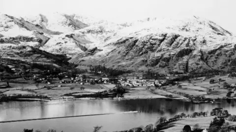 National Motor Museum/Heritage Images/Getty Images A black and white aerial shot showing Bluebird K7 racing across Coniston Water. Snow-capped mountains can seen seen in the background.