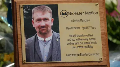 A gold plaque among the flower tributes. It includes an image of Dave Chester and a logo for Bicester Motion. It says: "We will cherish you Dave and you will be sorely missed, as we send our utmost love to Sue, Jordan and Riley. Love from the Bicester Community"
