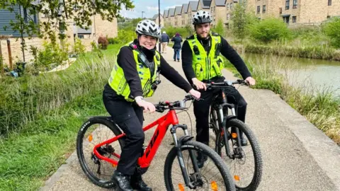 Wiltshire Police PC Huntington and PC Hawkins on bicycles on a footpath beside a canal with housing either side and lush green banks. They are both smiling at the camera in helmets and high-vis uniform.