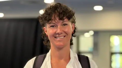 A woman with short brown curly hair wearing a white shirt, smiling at the camera