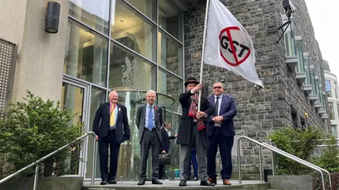 Four men stood outside of Guernsey's Royal Court. One is waving a big flag with a GST sign with a strikethrough on it. 