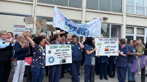 The picture shows NHS staff and members of the public outside the hospital. Some are holding placards.