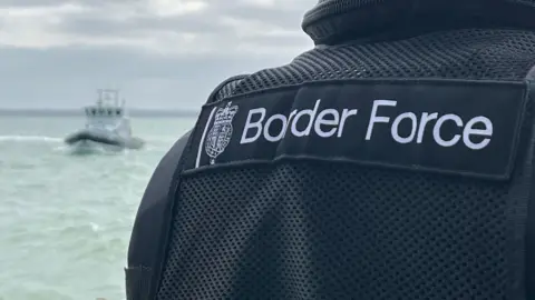 BBC Under grey skies, an anonymous uniformed officer from Border Force looks out to sea with a Border Force vessel in the distance. 