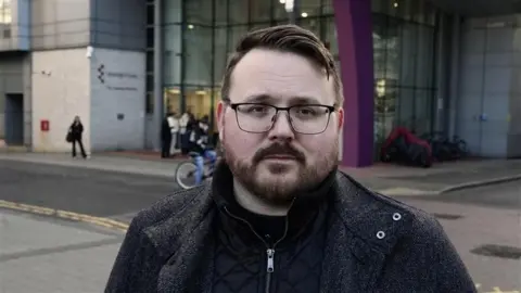 Stuart Woodward/BBC A man wearing a black quarter zip and a black coat. He has a beard and black frame glasses. He looks concerned and is standing in front of the Southend-on-Sea campus 