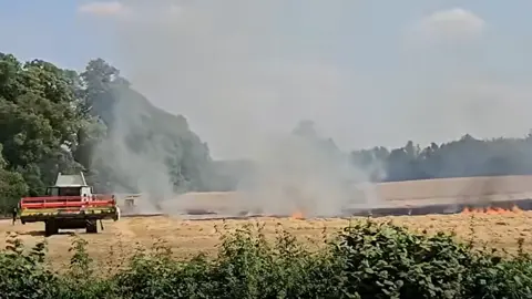 Richard Harvey A large field of straw affected by a large fire. Flames are low down with high plumes of smoke coming from the field. An area of the land is scorched. There is a farming vehicle on the left of the image which is not involved in fire. 