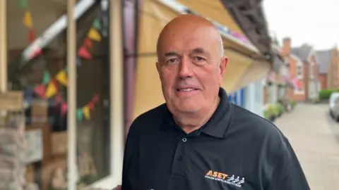 BBC A man in a dark polo shirt, with the words Audlem Special Events Team on it. He is stood outside a shop, and smiling at the camera