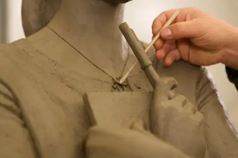 Discover Leicester Square A close-up of a sculpture of a woman being crafted, focusing on the neck, shoulders and chest. The sculpture is clutching a book in their hands across the chest. The sculptor is using a fine stick to craft the sculpture.