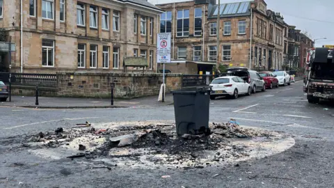 A bin in the middle of the street with the ground burned around it. The burnt area is all rubble and charcoal. 