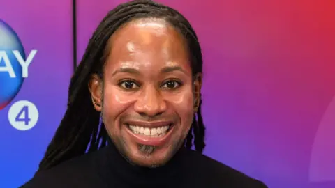 Prof Arday is smiling. He's wearing a black top and has earrings in. A backdrop with an ombre of colours is behind him. 