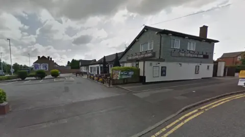 Google A two-storey pub site on the right with a car park and bushes on the left. Part of a road, including double yellow lines, is on the bottom right of the image.