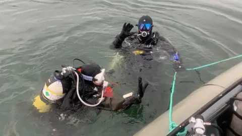 Two divers from Clean Planet UK in the ocean holding a green rope attached to a boat. One of the divers with blue glasses waves at the camera. 