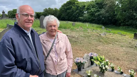 Nathan Turvey/BBC Pat Carey (right) who has white hair and is wearing a pink coat and her brother Peter Elton (left) who is wearing glasses and a blue coat, standing by the grave of the 15 children, there is a gravestone and flowers, with other gravestones and trees in thr distance. 