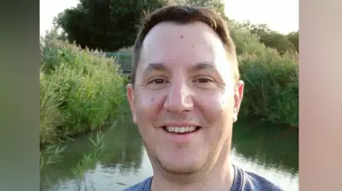 James Clements is standing in front of a small lake which is surrounded by tall grass. He has short brown hair, and is smiling at the camera. He is wearing a blue T-shirt.