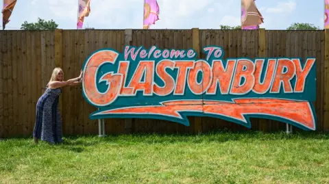 A woman posing next to a sign which says Welcome To Glastonbury