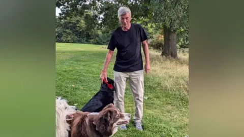 A picture of Graham Williams with three dogs, one white, one brown, and one black, standing in the park in front of a tree