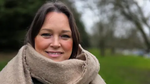 Paul Moseley/BBC Lucy Shires is standing on grass in front of some trees. She's smiling and wearing a large beige scarf and looking directly at the camera. 