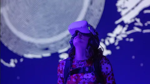 Mark McNulty Woman wearing a VR headset at the exhibition, gazing upwards in front of a blue and white exhibition display