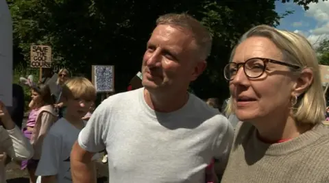 Charlie Caffyn wearing grey t-shirt and Emily Middleton wearing brown jumper stood with children behind them at protest.