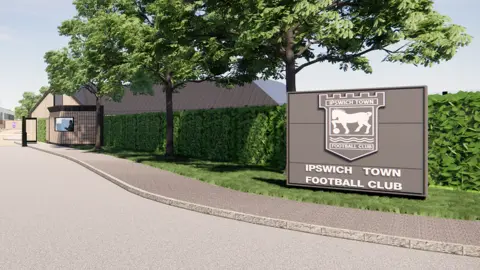 Ipswich Town Football Club Roadway onto site, with pavement and large "Ipswich Town Football Club" sign. There are trees and a hedge lining the route into the training ground. A security gatehouse is visible by the gate to the left of the picture.