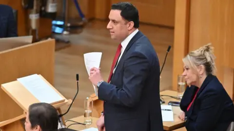 Getty Images Anas Sarwar, who has short black hair, stands up at a podium in the Scottish Parliament. He is wearing a dark suit, white shirt and red tie, and holding white papers . 