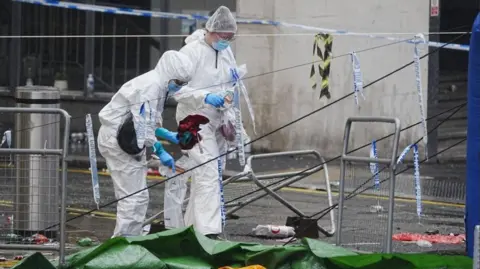 PA Media Forensic officers are at the scene in Water Street near the Liver Building in Liverpool. They are wearing while coveralls, face masks and hair coverings and are looking at the ground.