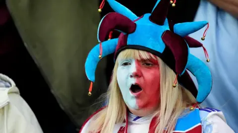 An Aston Villa fan in the stands before the UEFA Europa League match at Villa Park, Birmingham. She has long blonde hair and her face is painted half white, half red. She is wearing a maroon and blue jesters hat and her mouth is open as if mid chant or song.