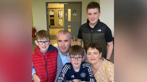 Fergal Sherry The Sherry Family pose for the camera while at Belfast City Hospital. There are two young boys sitting beside Fergal and Catherine Sherry, one is wearing a red gilet and the other a black Manchester United jersey. They both have medium-length light brown hair and are wearing dark, squared framed glasses. Fergal Sherry has greying hair and is wearing a light blue shirt. Catherine is sitting on the right and has short brown hair and is wearing makeup and a yellow, blue and pink floral shirt. Standing behind them is their oldest son, who is wearing a black and dark green North Face gilet.