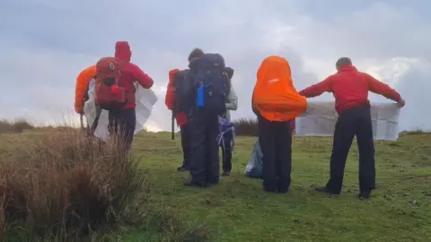 A group of teenagers stood holding a map as they try and navigate around on the moors. They are all wearing red rain jackets and big rucksacks on their backs.