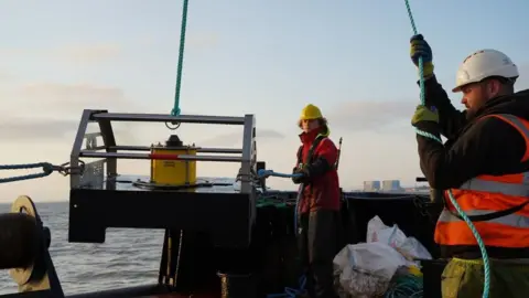 Two people in hard hats and high vis jackets stood near water with machinery they are pulling out of it. 