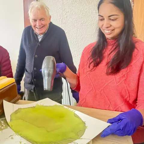 Young at Heart A man with short grey hair, light shirt, blue cardigan and grey trousers stands next to a woman who has long dark hair, a pink jumper and purple gloves. She is holding a hairdryer to dry green paint on a piece of card or paper.