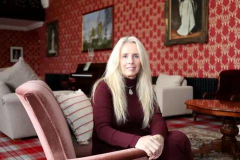 Peter Jolly/Northpix Suzanne has long blonde hair and is wearing a burgundy top. She is sitting in an armchair in a large room with ornately decorated red wallpaper.
