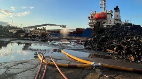 Humberside Fire and Rescue Service A long-armed aerial platform sprays water over the scene of the fire, with hoses in the foreground and a blue and white ship in the background