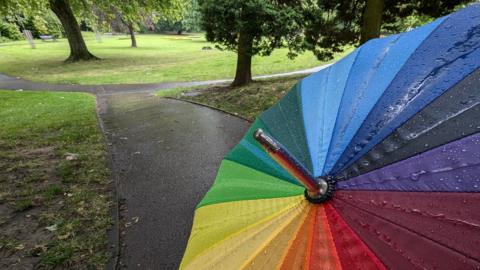 Multi-coloured umbrella with raindrops on. Wet park grounds in the distance. 