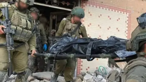 Israeli IDF soldiers with blurred faces carry a body in a black body bag, believed to be that of Yahya Sinwar, out of a destroyed house with a distinctive tiled roof on the front. 