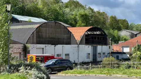 BBC Two buildings with metal arched roofs and white painted brick walls on the Fairfield industrial park. One appears to charred and a fire engine is stationed outside. Large trees grow behind the units and a few other industrial buildings are in the background.