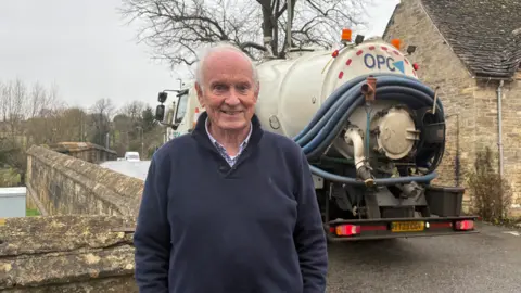Ken Gray stands by Burford bridge which has a tanker lorry crossing it. He has receding white hair and a dark blue top over a checked shirt.