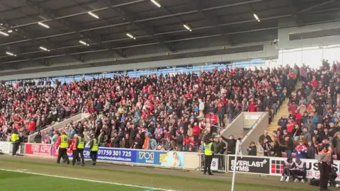 BBC/Richard Fox A group of mixed gender football fans celebrate a victory in the stands of a stadium.