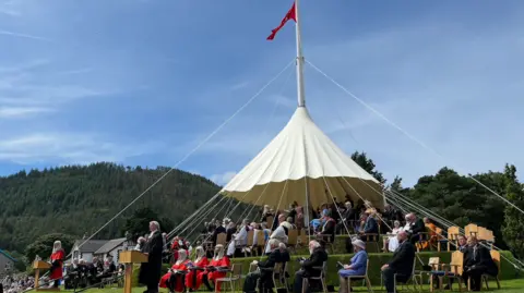 A large tent structure wrapping around a large flag pole. Lots of smartly dressed individuals are sitting in a circular formation around the tent. 