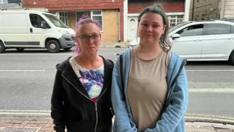 BBC Kelly Inch and her daughter Lou standing on a footpath with a car and van going past behind