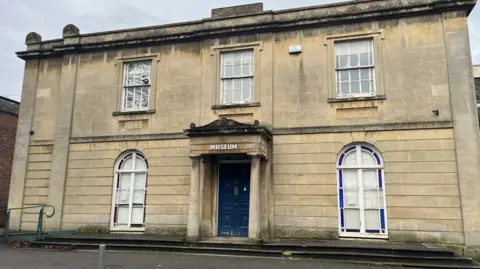 The exterior of Aplsey House. It is a large sand-coloured Georgian building, with two arched windows either side of the door and three rectangular sash windows upstairs. The blue front door has a porch overhang supported by two stone columns. The building appears somewhat run down with graffiti, a smashed window and a dirty façade.