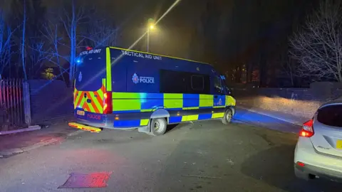 Greater Manchester Police A Tactical Aid Unit parked up on a road in Gorton. A streetlight is behind the van and there is a wall with trees to either side of the road. 